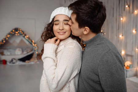 Young guy kissing her lovely girlfriend feeling happiness while celebrating winter holidays together, romantic couple hugging near decorated christmas tree in cozy home interior enjoying vacationsの写真素材
