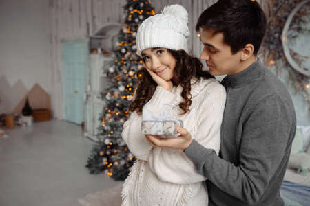 Man giving a Christmas present to his girlfriend. Lovely young couple hugging standing near decorated christmas tree celebrating Christmas at homeの写真素材
