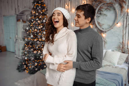 Cheerful couple laughing and enjoying family cozy moments indoors during New Year holidays, having fun together in cozy room with Christmas treeの写真素材
