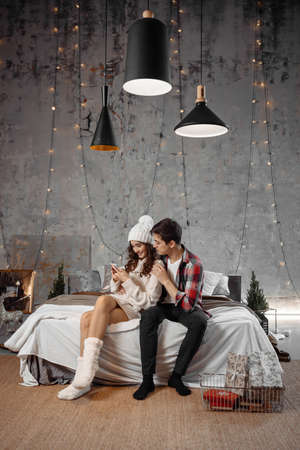 Full length photo of two cheerful people use cellphone man show x-mas seasonal discounts congratulations with Christmas sit on bed indoor with newyear decoration atmosphereの写真素材