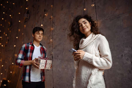 Cute young curly lady in white sweater using the phone while her boyfriend is getting ready to give her a present. Joyful cozy moments in winter holidays. Seasonal greetings. Advertising conceptの写真素材