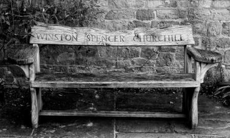 Wooden garden bench with the name Winston Spencer Churchill carved in the back from Chartwell House, Kent, his home until he died in 1965.  Afgainst a stone wall on stone flagging.  Black and white .のeditorial素材