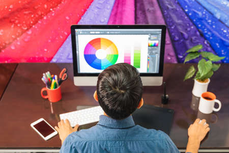 Back view. Young bearded businessman in denim shirt is sitting in office at table and is using computer with color swatch on screen. On table is smartphone and stationery. Man workingの写真素材