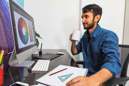 Side view. Smiling young bearded businessman in denim shirt is sitting in office at table. Drinking...