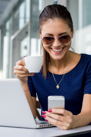 Excited beautiful girl reading e-mail with good news in a smartphone in a coffee shop with a window in the backgroundの写真素材