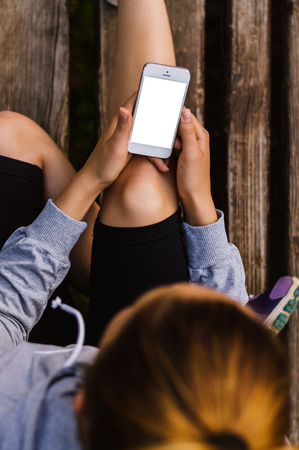 Woman using white smartphone while sitting on the bench. Blank screen, fitness enviromentの写真素材
