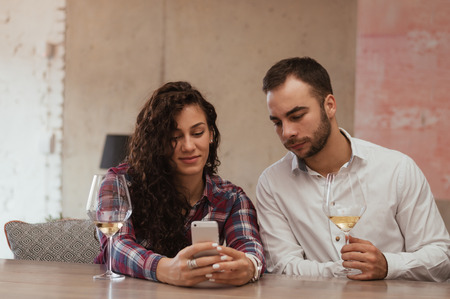 Couple drinking wine and looking at smartphone. In a restaurant cheerful couple surfing the web, looking a photo on smartphoneの写真素材