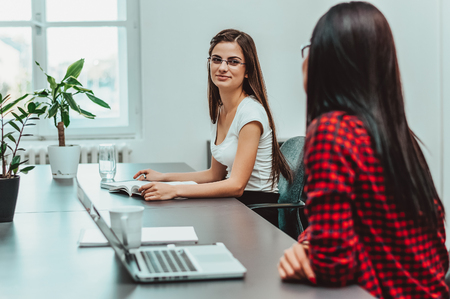 Happy young businesswomen working together at desk in modern office, looking at each other and talking.の写真素材