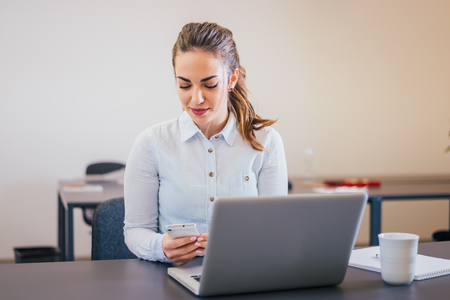 Beautiful young business woman looking at the phone while sitting in front of a laptopの写真素材
