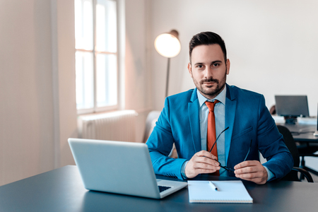 Portrait of young smiling businessman with laptop sitting in modern office.の写真素材
