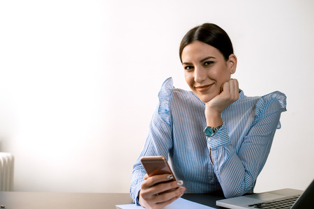 Woman in a blue shirt using phone while sitting at desk. Looking at camera.の写真素材