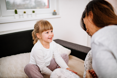 Adorable blonde little girl having fun with mother. Sitting in pajamas on the bed.の写真素材
