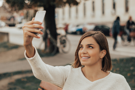 Young beautiful brunette taking pictures of herself on a cellphone in a city park.の写真素材