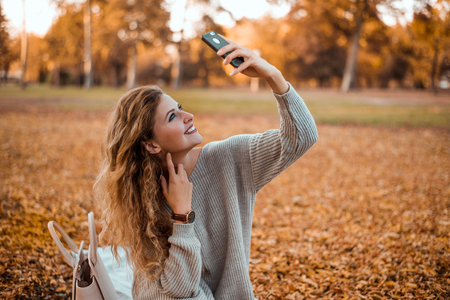 Beautiful blonde girl wearing modern autumn outfit sitting in park in autumn taking a selfie on smartphone.の写真素材