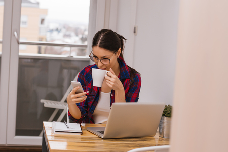 Girl in her appartment drinking coffee and using phoneの写真素材