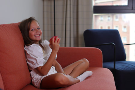 Smiling child girl sitting on sofa with red apple, healthy eatingの写真素材