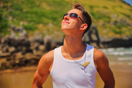 Young male in white sleeveless t-shirt and sunglasses on summer beach, tan skin, casual teenager lifestyle conceptの写真素材