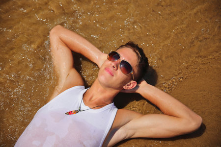 Young male in white sleeveless t-shirt and sunglasses on summer beach, tan skin, casual teenager lifestyle conceptの写真素材