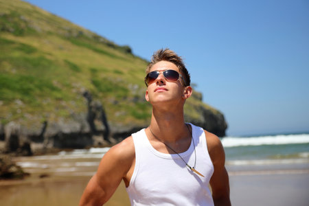 Young male in white sleeveless t-shirt and sunglasses on summer beach, tan skin, casual teenager lifestyle conceptの写真素材