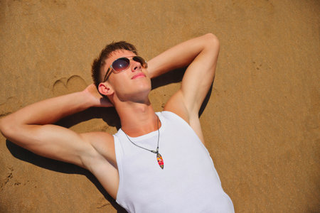 Young male in white sleeveless t-shirt and sunglasses on summer beach, tan skin, casual teenager lifestyle conceptの写真素材