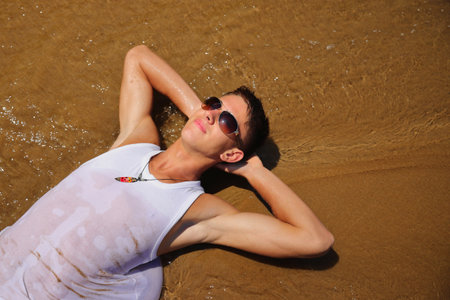 Young male in white sleeveless t-shirt and sunglasses on summer beach, tan skin, casual teenager lifestyle conceptの写真素材