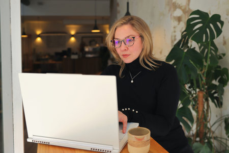 A woman in stylish glasses and a black turtleneck is engrossed in her smartphone while a laptop sits...