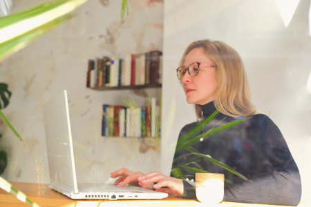 A woman in stylish glasses and a black turtleneck is engrossed in her smartphone while a laptop sits...
