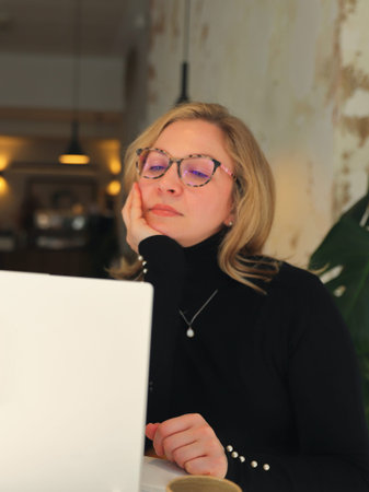 A woman in stylish glasses and a black turtleneck is engrossed in her smartphone while a laptop sits...