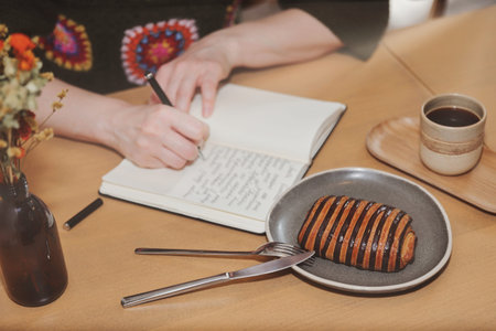 A woman with blonde hair wearing a crochet sweater writes in a journal at a cafe table, with a...