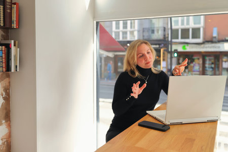 A woman in stylish glasses and a black turtleneck is engrossed in her smartphone while a laptop sits...