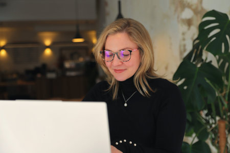 A woman in stylish glasses and a black turtleneck is engrossed in her smartphone while a laptop sits...
