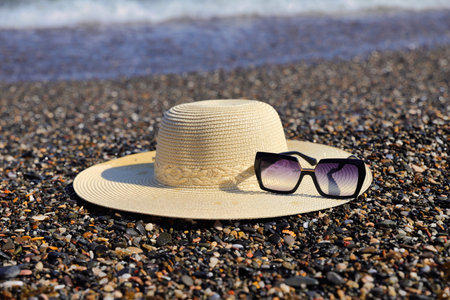 A wide-brimmed straw hat and stylish sunglasses are placed on a wet pebble beach, with the ocean's edge visible in the distance