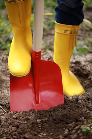 A woman in yellow boots is digging soil with a shovel in a gardenの写真素材