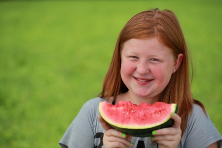 Red-haired girl eating a watermelon on a green meadow. Blurred background.の写真素材