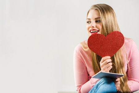 Young pretty smiling blonde girl with long hair holding tablet and heart in hands and dreaming abount something. White background. Valentine's Day, Christmas, Birthday, Holidays concept.の写真素材