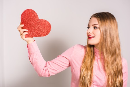 Portrait of a young smiling blonde girl with red lips wearing shirt and holding heart in her hands. White background. Valentine's Day, Christmas, Birthday, Holidays.の写真素材