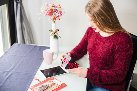 Young pretty smiling blonde girl with maroon lips and sweater sitting at the table and opening a...
