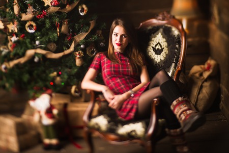 Young cute happy girl in red dress sitting in chair close to Christmas tree and gifts on wooden background. New Year and Christmas celebrationの写真素材