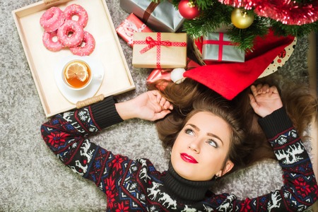 Young cute happy girl in sweater with deer lying on the floor at home on Christmas. New Year and Christmas celebration. Christmas tree, gifts, tea and sweets on backgroundの写真素材