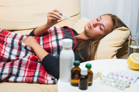 Cute girl caught cold. Sick woman lying on sofa under wool blanket and looking at thermometer. Illness concept. Pills, lemon and glass of water on tableの写真素材