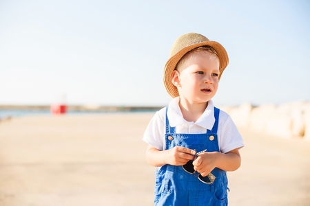 Portrait of a cute small blond boy with blue eyes in a blue overall, white shirt and hat holding glasses on a beach in summerの写真素材