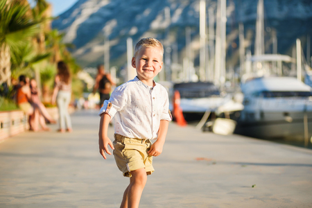 Happy cute small blond boy in white shirt smiling and playing in port with yachts on backgroundの写真素材