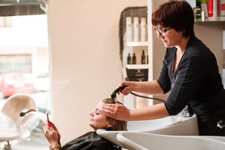 Hairdresser and a woman in a beauty salon during a hair wash. Woman with a mobile phone.の写真素材