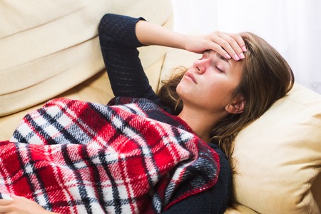 Cute girl caught cold. Sick woman with red nose lying on sofa under wool blanket and holding hand on headの写真素材