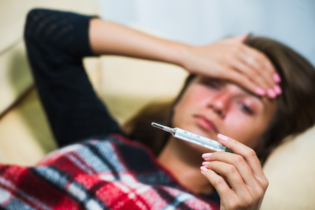 Cute girl caught cold. Sick woman with red nose lying on sofa under wool blanket, holding hand on head and looking at thermometer. Illness conceptの写真素材