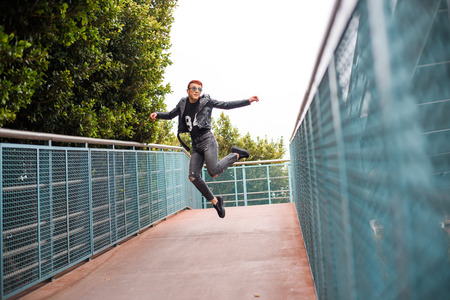 Young stylish boy wearing sunglasses, leather jacket and ripped jeans is jumping on a bridge on an...