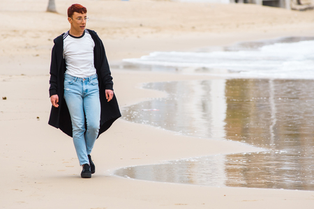 Young stylish male gay with red hair wearing glasses, long sport jacket and blue jeans is walking on a beach.の写真素材