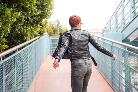 Young stylish boy wearing sunglasses, leather jacket and ripped jeans is turning around on a bridge...
