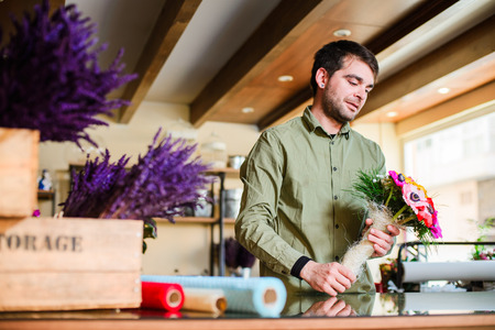 Male attractive smiling florist wearing green shirt and making bouquet in a flower shop. Man assistant or owner in floral design studio, making decorations. Flowers delivery, creating orderの写真素材
