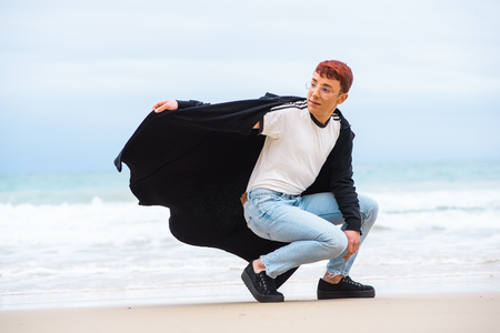 Young stylish male gay with red hair wearing glasses, long sport jacket and blue jeans is posing on a beach.の写真素材